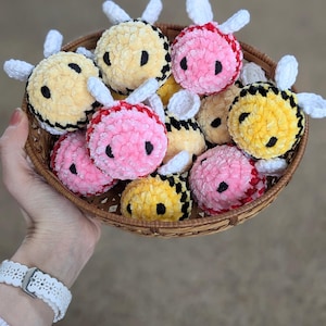 May include: A basket filled with crocheted bee plushies. The bees are yellow, pink, and black with white wings.