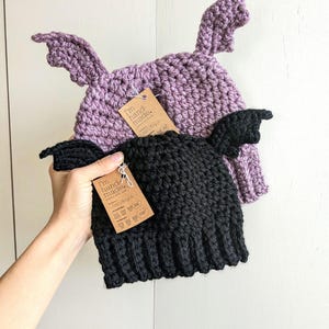 May include: Two crocheted beanies, one in lavender and one in black, each featuring two small bat-like wings on the sides. Both hats have a brown cardboard tag attached that reads I'm hand-made with care and includes various laundry care symbols.