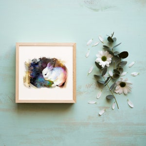 May include: A square wooden frame containing a print of a white rabbit set against a dark, colorful, galaxy-like background. The rabbit is depicted in a painterly style with soft edges, positioned next to a sprig of eucalyptus and white flowers.