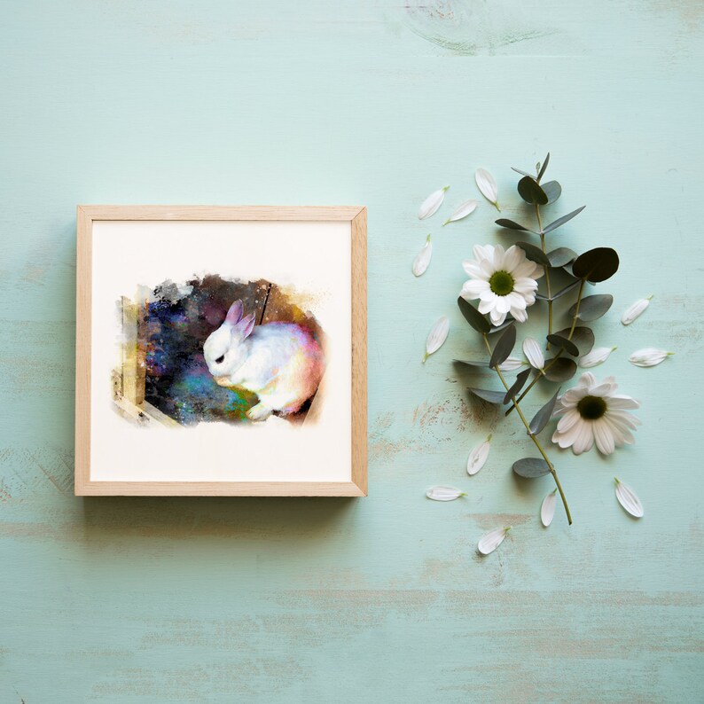 May include: A square wooden frame containing a print of a white rabbit set against a dark, colorful, galaxy-like background. The rabbit is depicted in a painterly style with soft edges, positioned next to a sprig of eucalyptus and white flowers.