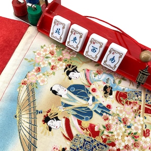 May include: A red Mahjong set with white tiles featuring Chinese characters. The set includes a red tray, colourful chips, and a decorative fabric with an Asian-inspired illustration of people and cherry blossoms.