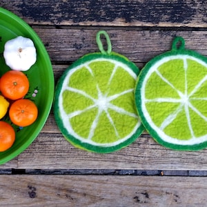 May include: A green tray displays tomatoes, tangerines, a lemon, garlic, and a cup of liquid. Two lime-shaped felt trivets with green and white segments are placed nearby. The scene is on a wooden surface, with a glimpse of green shoes.