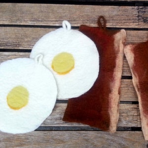 Könnte beinhalten: Zwei weiße Filz-Topflappen in Form von Spiegeleiern, zwei braune Filz-Topflappen in Form von Speckscheiben und ein Teller mit zwei gelben Bananen, einer Orange und einer Kiwi.