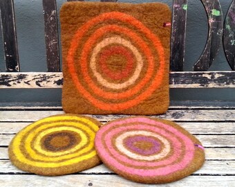 May include: Three round felt trivets with concentric circles in different colours. The trivets are brown, yellow, and pink with brown, orange, and purple circles.