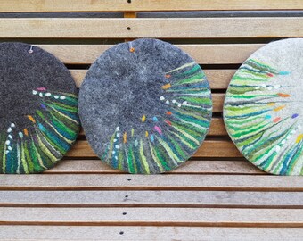 May include: Three round felt coasters with a green, white, and multi-colored design. The coasters are arranged in a row on a wooden surface.