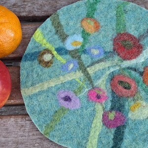 Op de afbeelding: Een stilleven met een rode appel, een sinaasappel en een ronde, turquoise vilten onderzetter. De onderzetter heeft een bloemenontwerp in verschillende kleuren. De vruchten liggen op een houten oppervlak. De onderzetter heeft een diameter van ongeveer 20 cm.
