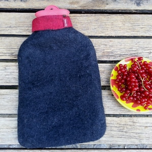 以下が含まれることがあります： 赤いフェルトのトップとピンクのプラスチック製の蓋が付いた黒いフェルトの湯たんぽ。湯たんぽは、赤いカラントの入った黄色と赤のプレートの隣の木製表面の上に置かれています。