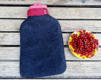 May include: A black felt hot water bottle with a red felt top and a pink plastic lid. The bottle is sitting on a wooden surface next to a yellow and red plate with red currants.