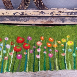 Könnte beinhalten: Ein grüner Filz-Wandbehang mit einem farbenfrohen Blumendesign. Die Blumen sind in Rot-, Orange-, Gelb-, Rosa- und Blautönen gehalten. Die Blumen sind in einer Reihe am unteren Rand des Wandbehangs angeordnet.