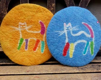May include: Two round felt coasters with a white cat design. The cat on the yellow coaster has a rainbow tail and legs. The cat on the blue coaster has a pink tail and legs.