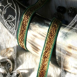 May include: A close-up of a decorative drinking horn with a woven ribbon. The ribbon is green and gold with a geometric pattern. The horn is light gray and brown. Several silver rings are visible.