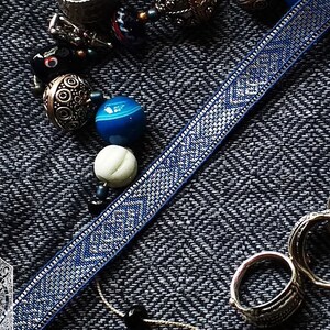 May include: An assortment of jewelry and accessories, including rings, beads, and a blue woven ribbon with a geometric pattern. A gold-colored Thor's hammer pendant is also visible. The text "Woven by Alicja Jaczewska" is at the bottom.