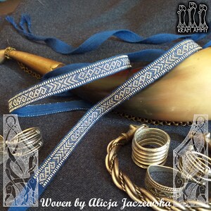 May include: A blue woven ribbon with a geometric pattern in white, displayed with silver rings and a brass horn. The text "Woven by Alicja Jaczewska" is visible at the bottom of the image.