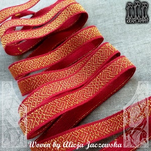 Può includere: Primo piano di un nastro tessuto rosso e oro con un motivo geometrico. Il nastro è piegato e stratificato, mostrando il design dettagliato. Il testo "Woven by Alicja Jaczewska" è visibile. Questo nastro è probabilmente utilizzato per l'artigianato o la decorazione.