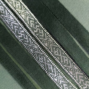 May include: Close-up of two silver and black patterned ribbons with a geometric design. The ribbons are placed on a dark green fabric background. The image shows the texture and detail of the ribbons.