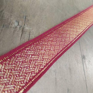 May include: A length of decorative ribbon with a woven gold pattern on a deep red background. The ribbon's design features a repeating geometric pattern, adding a touch of elegance. Ideal for crafting or embellishing various projects.