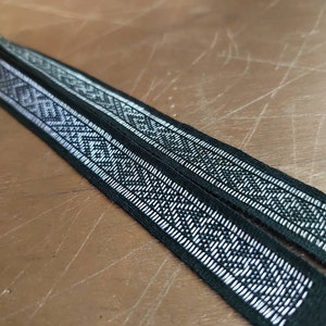 May include: Two black woven ribbons with a repeating geometric pattern in white. The ribbons are lying side by side, angled across a wooden surface. The detailed design is visible, showcasing the craftsmanship of the textile.