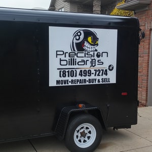 May include: A black trailer with a white sign that says "Precision Billiards (810) 499-7274 MOVE-REPAIR-BUY & SELL". The sign also features a cartoon 8-ball with a menacing expression.