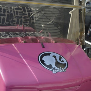 May include: A pink golf cart with a black and white patterned seat and a Barbie logo on the bonnet.