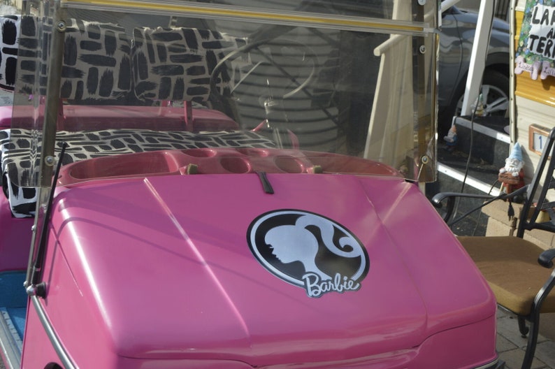May include: A pink golf cart with a black and white patterned seat and a Barbie logo on the bonnet.