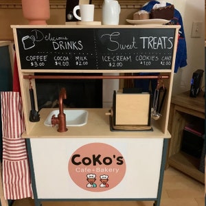May include: A wooden play kitchen with a black chalkboard sign that says "Delicious Drinks" and "Sweet Treats". The sign lists prices for coffee, cocoa, milk, ice cream, cookies, and cake. The kitchen has a sink, a toaster, and a sign that says "Coko's Cafe + Bakery".