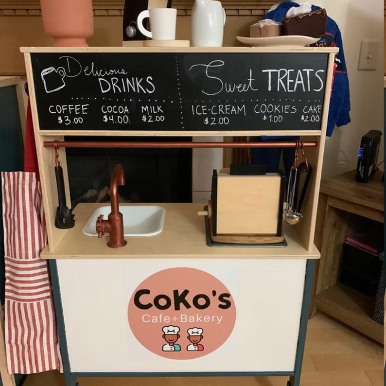 May include: A wooden play kitchen with a black chalkboard sign that says "Delicious Drinks" and "Sweet Treats". The sign lists prices for coffee, cocoa, milk, ice cream, cookies, and cake. The kitchen has a sink, a toaster, and a sign that says "Coko's Cafe + Bakery".