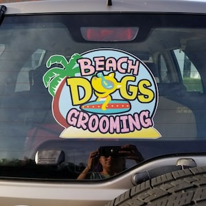 May include: A sticker on the rear window of a car that reads "Beach Dogs Grooming" with a cartoon dog wearing sunglasses and a surfboard. The sticker is on a blue background with a palm tree and a pink beach.