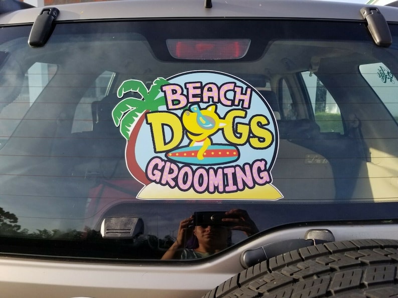 May include: A sticker on the rear window of a car that reads "Beach Dogs Grooming" with a cartoon dog wearing sunglasses and a surfboard. The sticker is on a blue background with a palm tree and a pink beach.