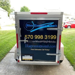 May include: A white trailer with a black and blue logo that says "Blue Pressure Wash" and a phone number 570 998 3199. The trailer also has the text "Concrete Wood Vinyl Bricks" and the website "Bluepressurewash.com". The trailer has a number plate that says "CARN'S TRAILERS".