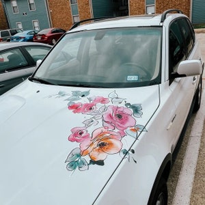 May include: A white SUV with a floral design painted on the bonnet. The design features pink, orange, and green flowers with black outlines. The date "02-22" is visible on the windscreen.