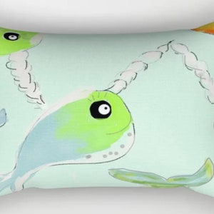 May include: Rectangular pillow with a whimsical design featuring cartoon narwhals in shades of green and blue, connected by white rope-like lines against a light green background. The narwhals have large black eyes and are smiling.