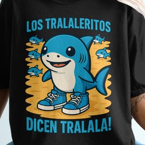 May include: Black t-shirt featuring a cartoon shark wearing blue sneakers. The shark is surrounded by small blue fish and the text "LOS TRALALERITOS DICEN TRALALA!" in blue. The shirt is worn by a person.