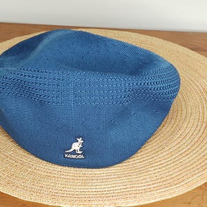 May include: A blue Kangol cap with a textured knit design. The cap features the Kangol logo on the front and is set against a straw hat on a wooden surface. The cap is a classic style.