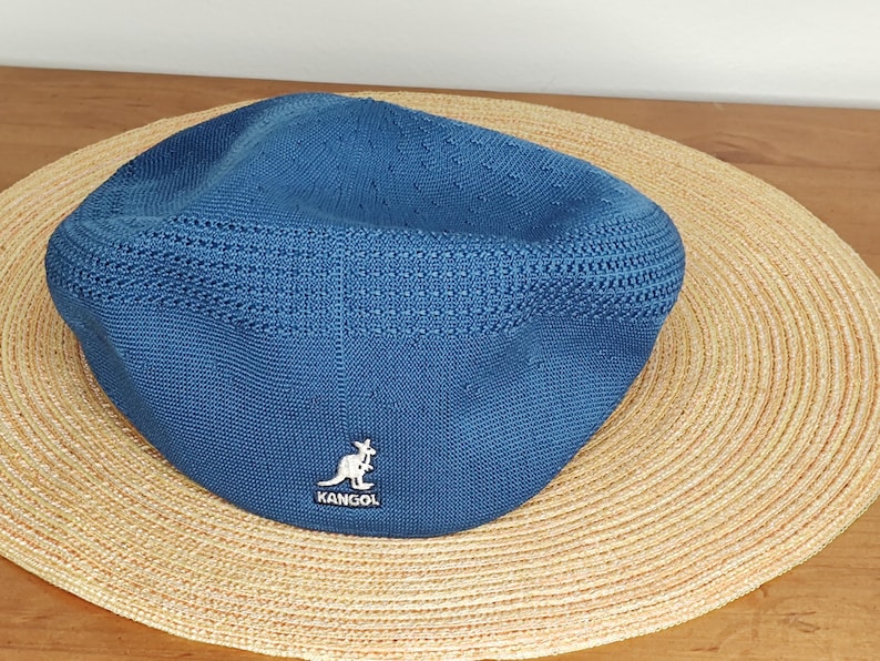 May include: A blue Kangol cap with a textured knit design. The cap features the Kangol logo on the front and is set against a straw hat on a wooden surface. The cap is a classic style.