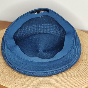 May include: A blue knit cap with a rounded crown and a short brim, set atop a wide-brimmed straw hat. The cap's interior is visible, revealing a darker blue lining. The hats are on a light brown wooden surface.