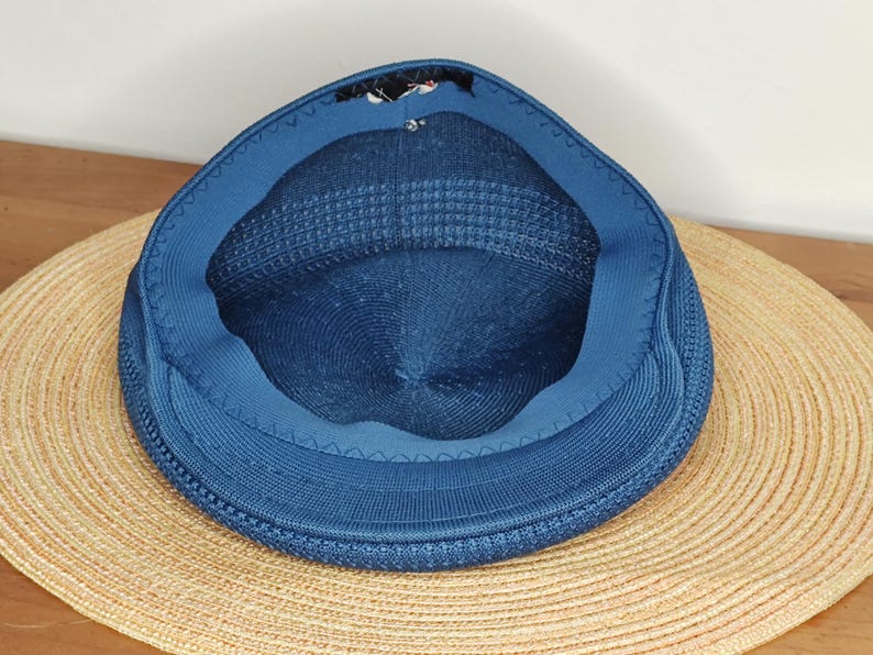 May include: A blue knit cap with a rounded crown and a short brim, set atop a wide-brimmed straw hat. The cap's interior is visible, revealing a darker blue lining. The hats are on a light brown wooden surface.