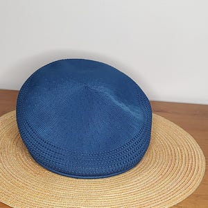 May include: A blue flat cap sits atop a wide-brimmed straw hat. The cap has a textured, woven appearance. The straw hat is circular with a woven pattern. The items are on a wooden surface.