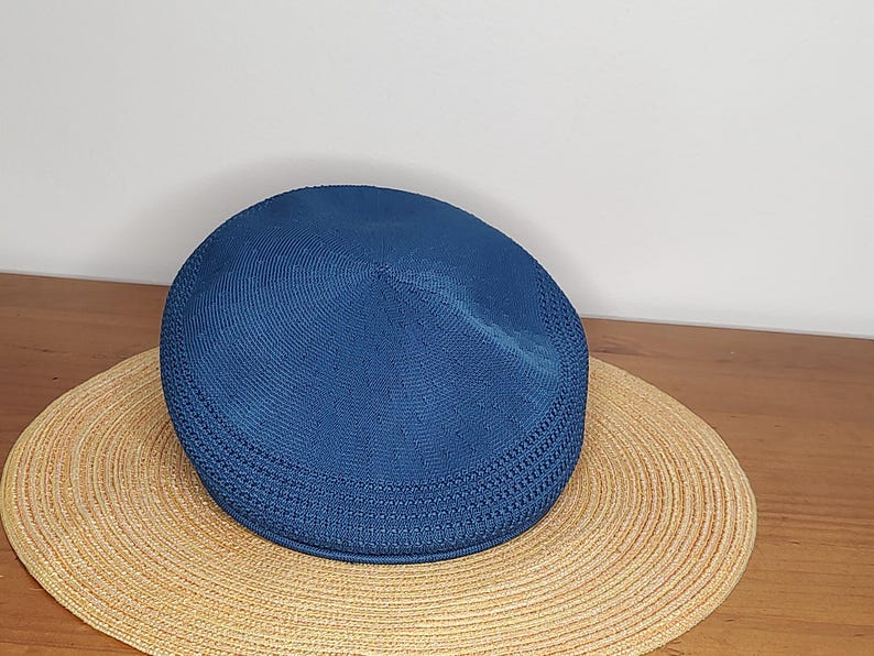 May include: A blue flat cap sits atop a wide-brimmed straw hat. The cap has a textured, woven appearance. The straw hat is circular with a woven pattern. The items are on a wooden surface.