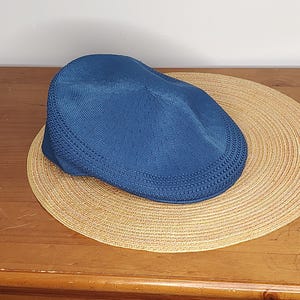 May include: A blue newsboy cap rests on a round, woven straw hat. The cap has a textured knit design. The straw hat is a light tan color and has a woven pattern. The items are on a wooden surface.