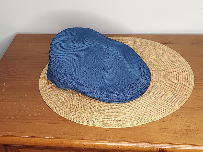 May include: A blue newsboy cap rests on a round, woven straw hat. The cap has a textured knit design. The straw hat is a light tan color and has a woven pattern. The items are on a wooden surface.