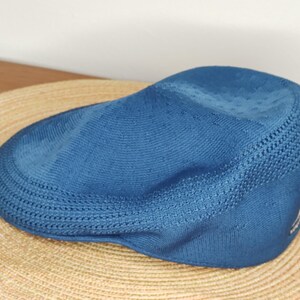 May include: A blue newsboy cap with a textured knit design. The cap is resting on a woven straw hat. The cap has a small logo on the side. The hat is a stylish accessory.