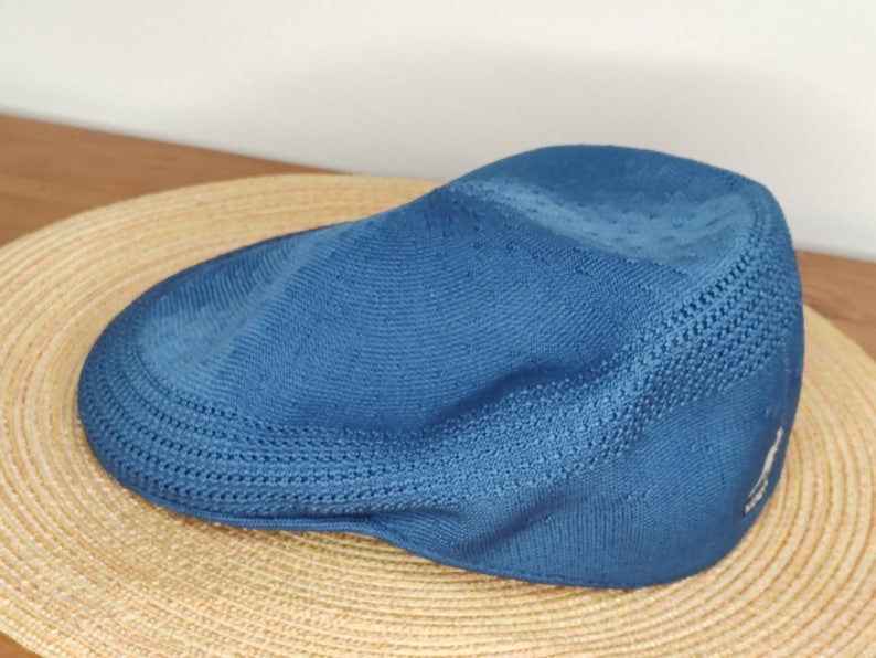 May include: A blue newsboy cap with a textured knit design. The cap is resting on a woven straw hat. The cap has a small logo on the side. The hat is a stylish accessory.