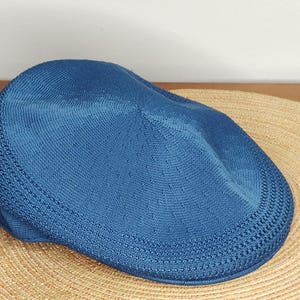 May include: A blue flat cap made of a knitted fabric. The cap is resting on a woven straw hat. The cap has a rounded crown and a short, stiff brim. The cap is a solid blue color.
