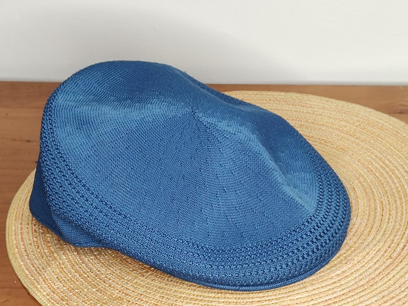 May include: A blue flat cap made of a knitted fabric. The cap is resting on a woven straw hat. The cap has a rounded crown and a short, stiff brim. The cap is a solid blue color.