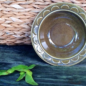 May include: A green ceramic bowl with a scalloped rim. The bowl is sitting on a wooden surface with green leaves.