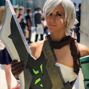 May include: A person in cosplay, holding a large, stylized sword. The sword is mainly grey and black with green accents. The person is wearing a white top, brown scarf, and a brown glove. The background is blurred, suggesting an outdoor event.