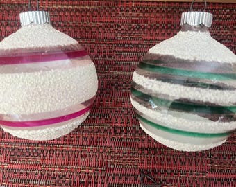 Könnte beinhalten: Zwei kugelförmige Glasornamente mit abwechselnden horizontalen Streifen aus klarem Glas und weißem, texturiertem Glitzer. Ein Ornament zeigt leuchtend magentafarbene Streifen, das andere dunkelgrüne Streifen. Beide haben silberne Metallkappen.
