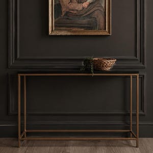 May include: A rectangular framed oil painting depicting an elderly person in a dark cap and brown coat reading a newspaper, positioned above a console table with a thin gold metal frame, a black top, and a small woven basket with greenery.
