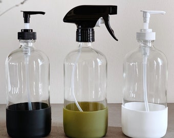 May include: Three glass bottles with different colored silicone bases. The bottles are clear glass and have different tops: a pump, a spray nozzle, and a pump. The bases are black, green, and white.