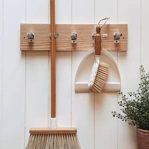 May include: A light-coloured wooden wall-mounted rack featuring five silver metal clips. A broom with a long wooden handle and a dustpan set with a wooden handle and a white marble-patterned brush are suspended from the clips on the wooden board.